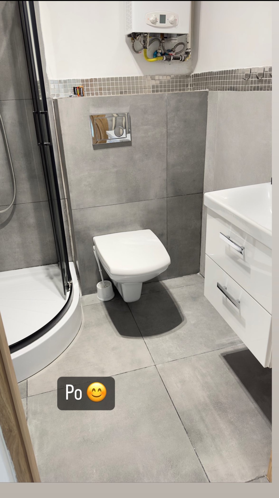 Wyremontowana łazienka z szarą glazurą, wiszącą toaletą, kabiną prysznicową z czarnymi ramami i białym zlewem z szafkami, widoczny bojler nad mozaiką.