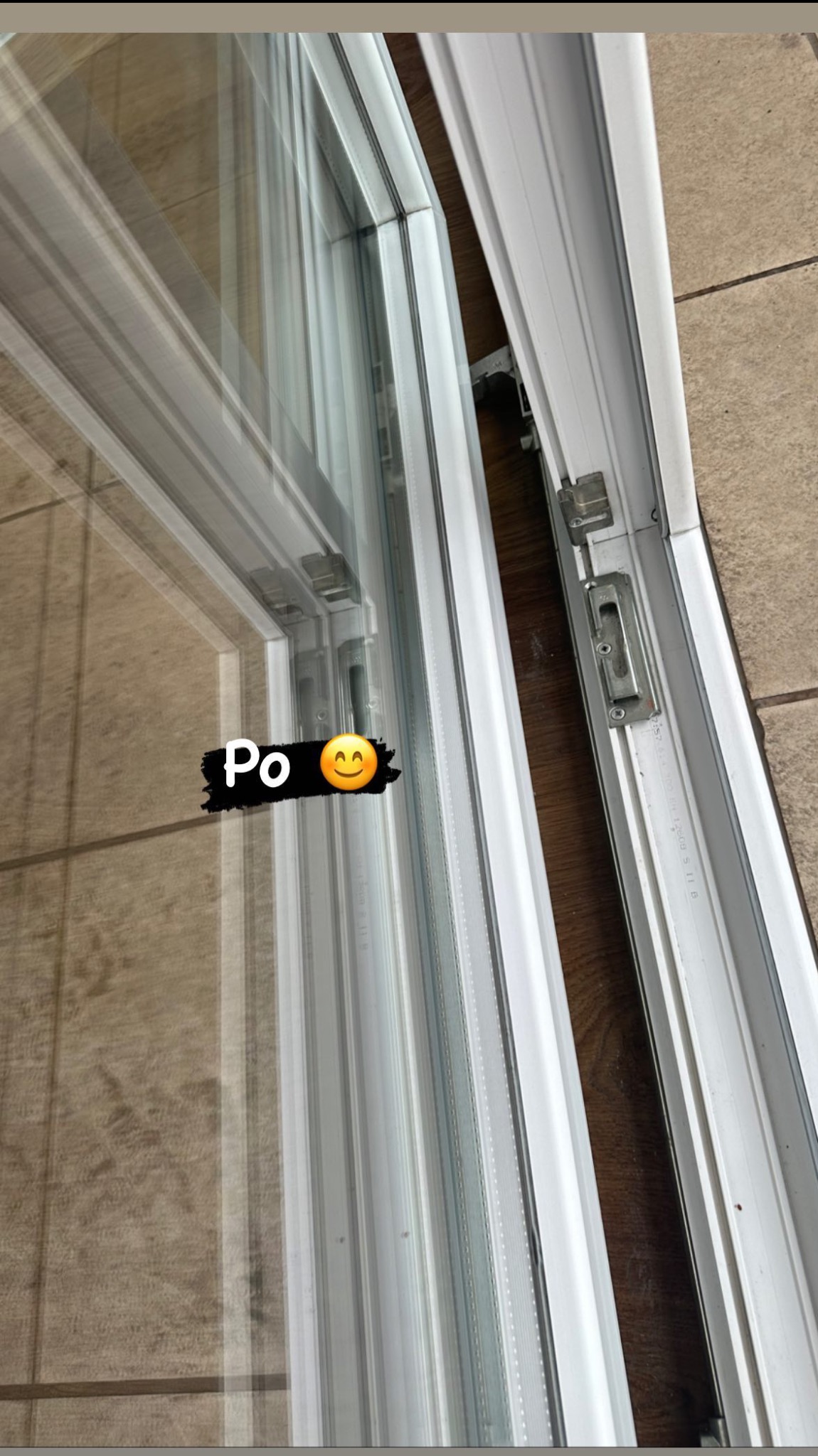 Błyszcząca, czysta szyba okienna z białymi ramami, widoczne okucia i mechanizm otwierania, odbicie podłogi w szybie.