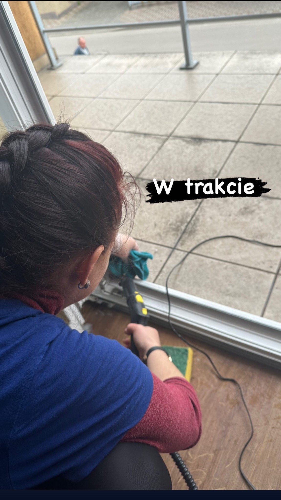 Kobieta z warkoczem czyści ramę okienną parownicą i szmatką, widoczny fragment podłogi z płytek na balkonie i oddalona postać mężczyzny.