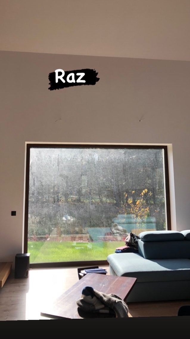 Duże okno w salonie z wyraźnymi smugami brudu i zaciekami, widok na zielony ogród, część sofy i stolika kawowego