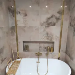 Nowoczesna łazienka z owalną białą wanną, złotą armaturą i marmurkowymi płytkami z akcentami w kolorze złota, p&oacute;łka w ścianie z dekoracjami, drewniana p&oacute;łka na wannę.