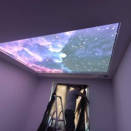 MARIUSZ STRZELAK - Montaż podświetlanego sufitu z motywem kosmosu: mężczyzna na drabinie, widoczne narzędzia i fragment okna z zasłoną.