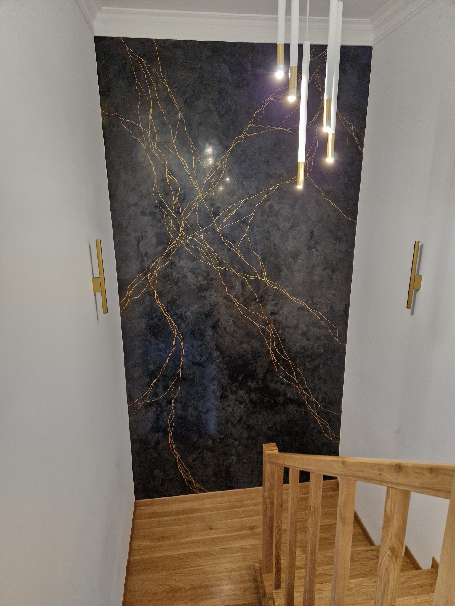 Ściana w klatce schodowej z dekoracyjną, ciemną gładzią imitującą marmur z nieregularnymi, złotymi żyłkami, oświetlona wiszącą lampą z białymi i złotymi elementami oraz złotymi kinkietami ściennymi.