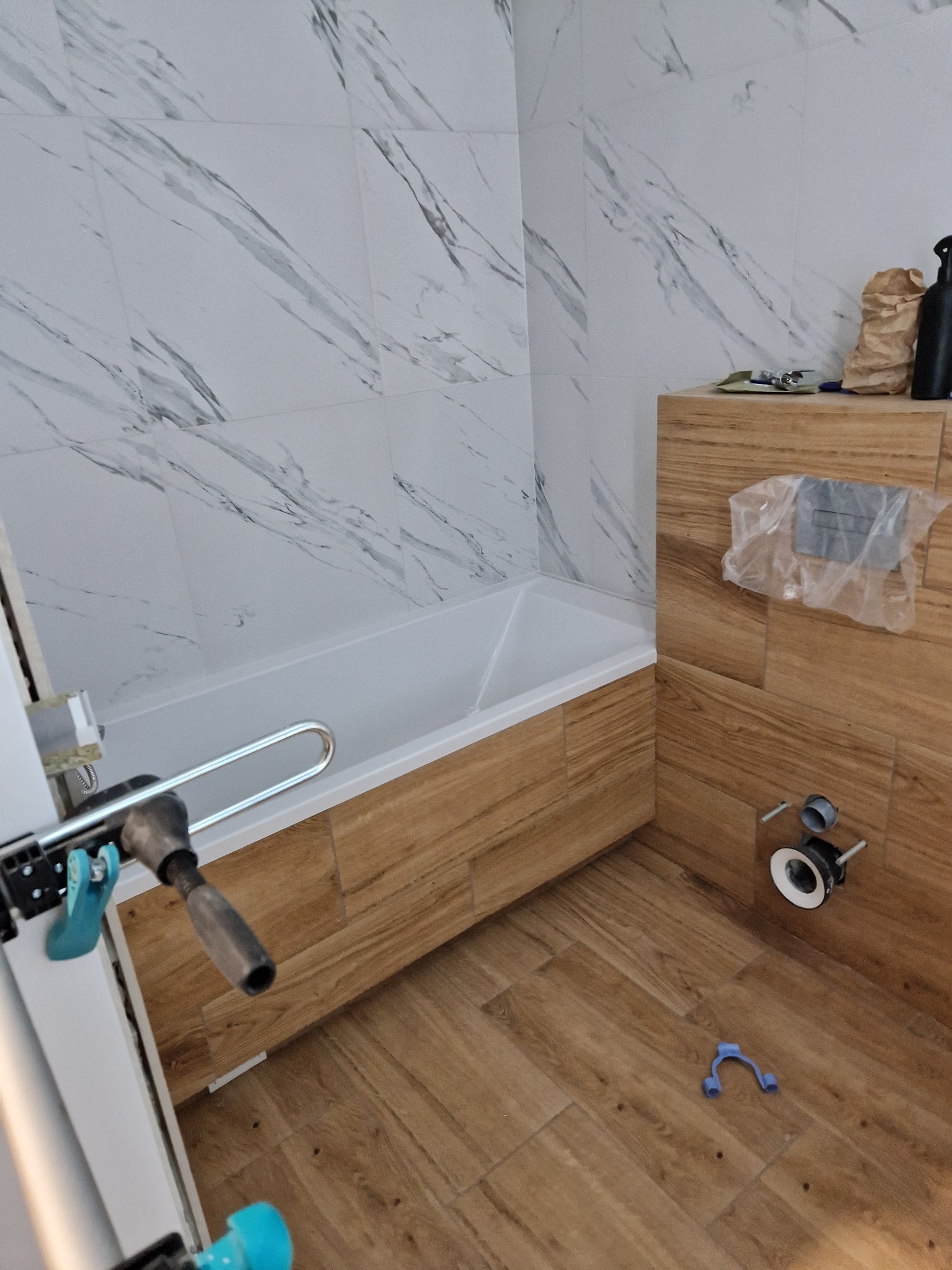 Wnętrze łazienki w trakcie remontu: widoczna wanna obudowana drewnopodobnymi płytkami, ściany wyłożone marmurowymi płytkami, elementy instalacji sanitarnej oraz narzędzia glazurnicze.