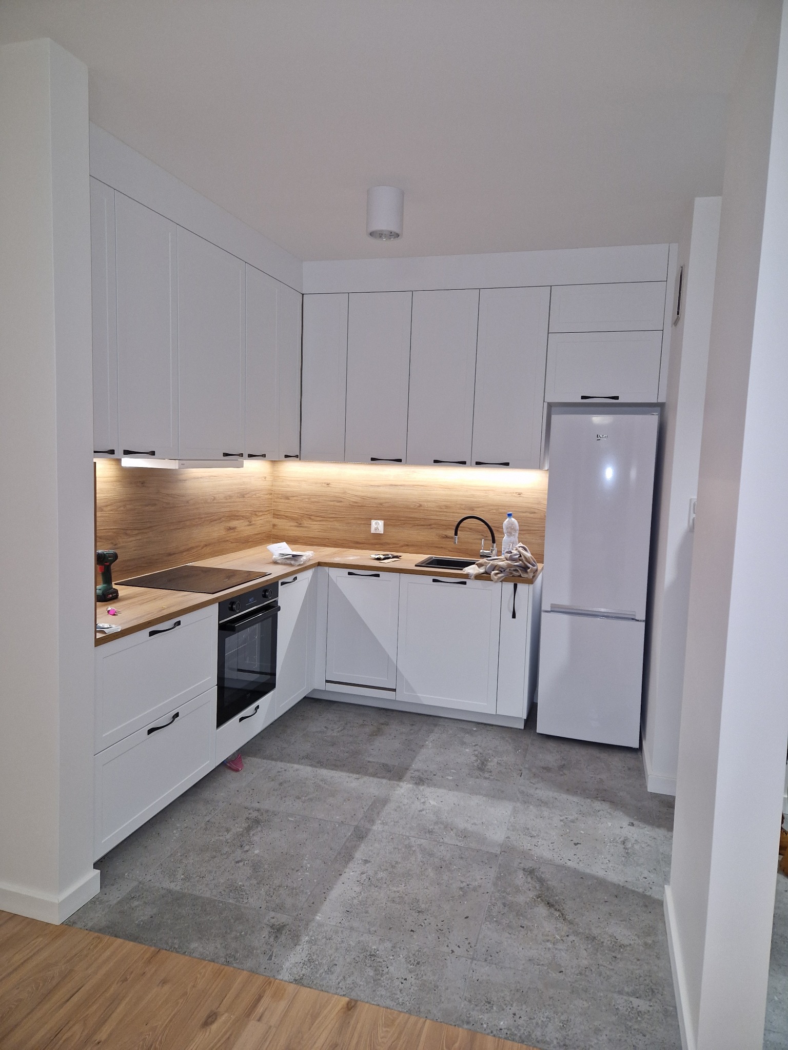 Kompaktowa kuchnia w stylu skandynawskim z białymi frontami szafek, drewnianym blatem i panelem ściennym imitującym drewno, szarymi płytkami podłogowymi i sprzętem AGD w zabudowie.