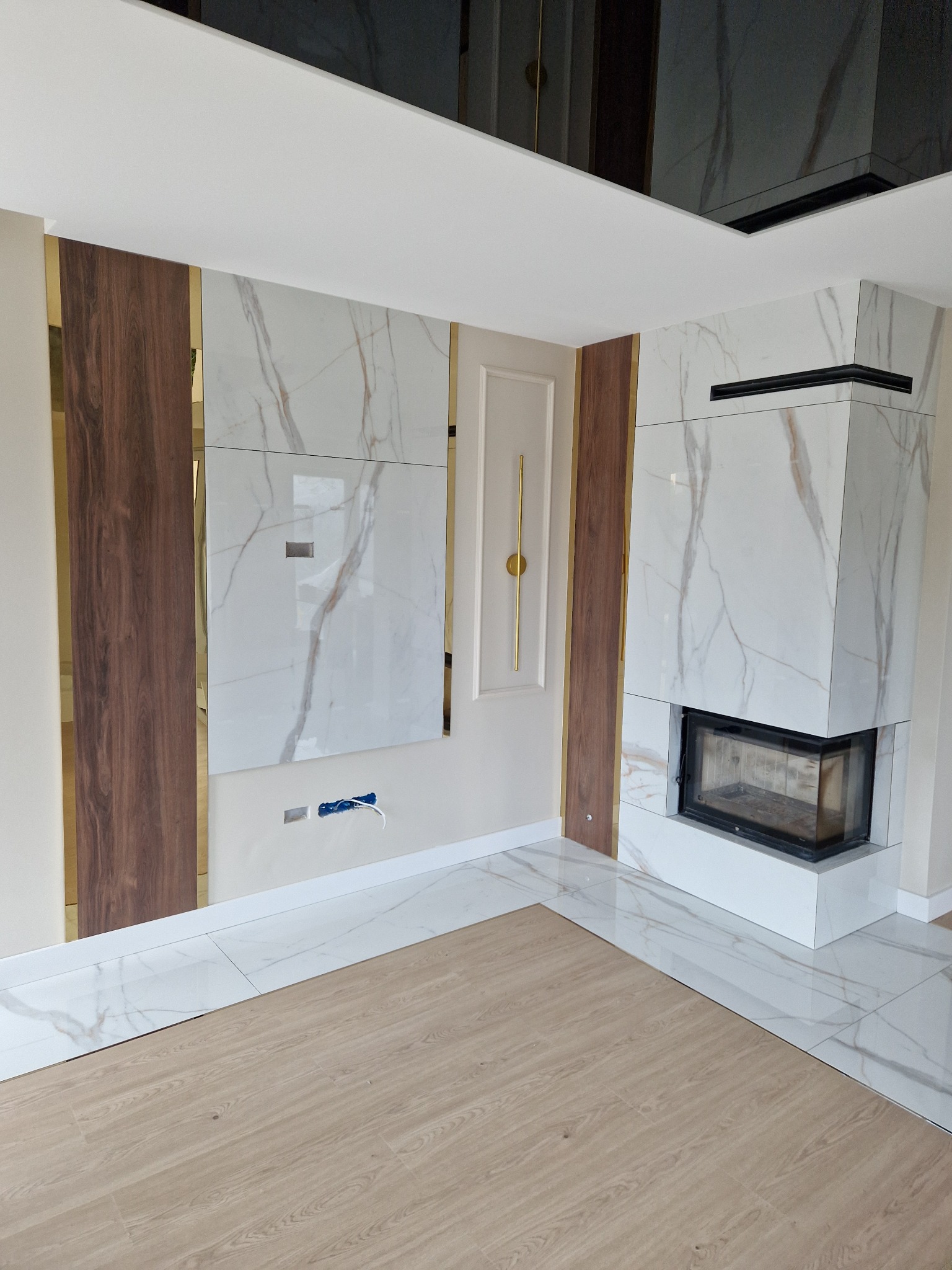 Nowoczesny salon z marmurową ścianą akcentującą kominek, drewnianymi panelami z lustrzanymi wstawkami i błyszczącym sufitem, częściowo wykończony parkiet i płytki podłogowe.