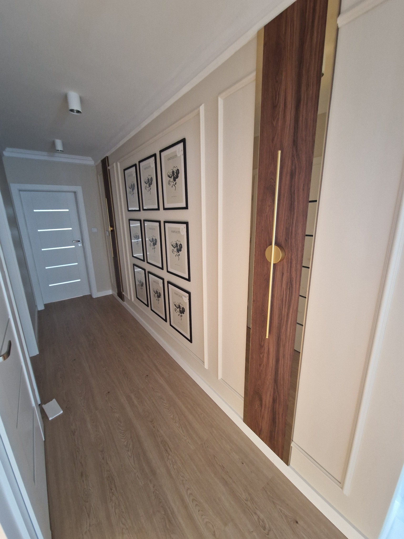 Nowoczesny korytarz z panelami ściennymi, drewnianą okładziną z lustrzanym paskiem i złotym uchwytem, parkietem oraz galerią czarno-białych grafik w ramkach.