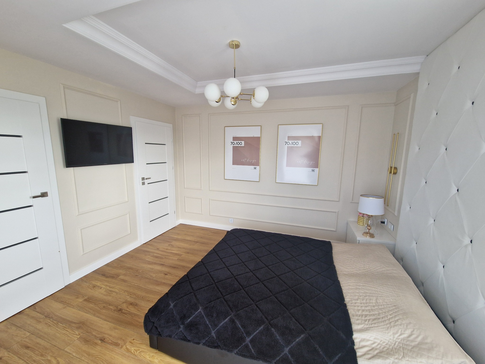 Elegancka sypialnia z beżowymi ścianami zdobionymi sztukaterią, drewnianą podłogą, łóżkiem z pikowanym zagłówkiem i czarną narzutą, nowoczesną lampą sufitową i dwoma plakatami w złotych ramach.