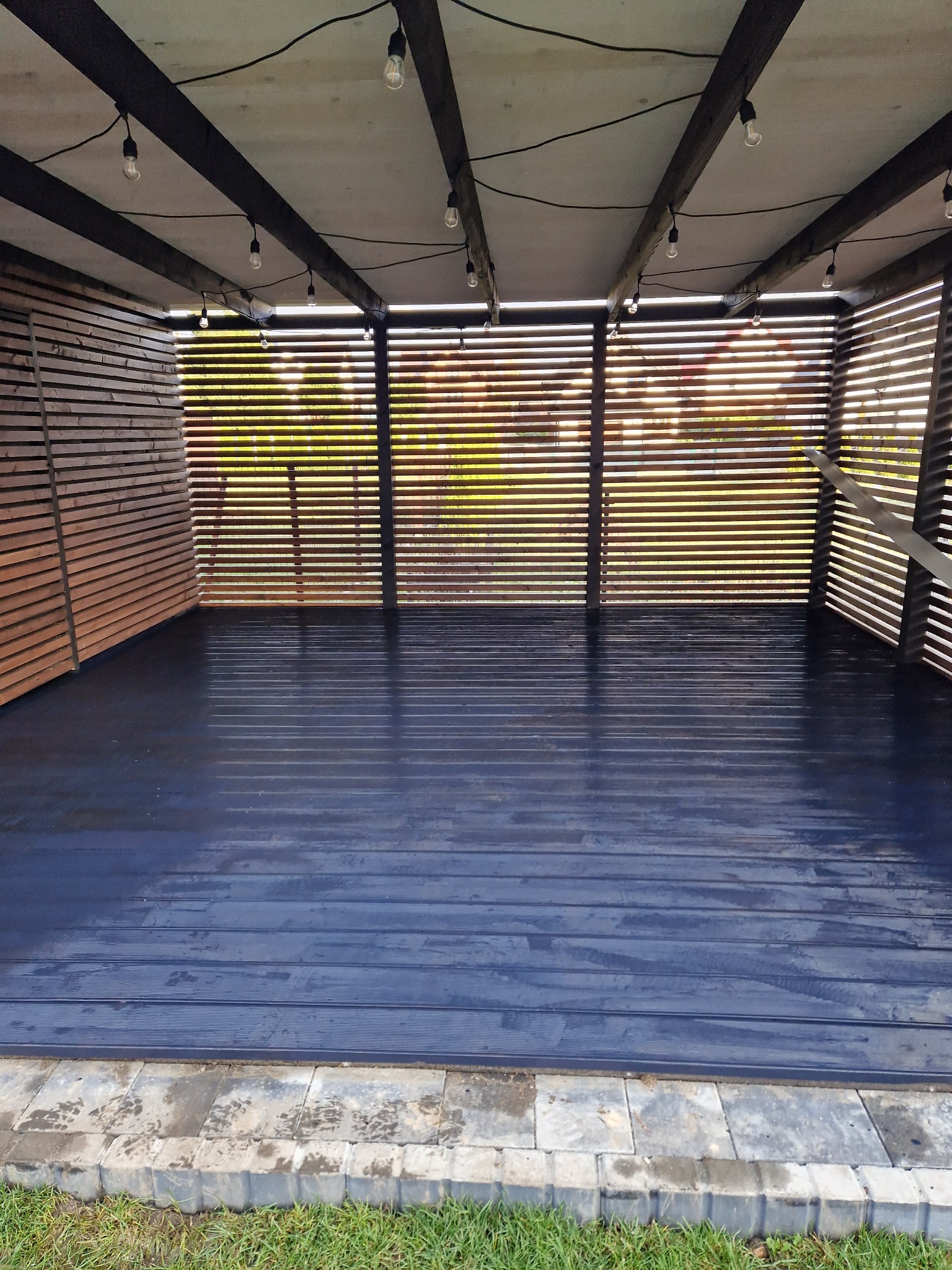 Zadaszony taras z drewnianą podłogą pomalowaną na ciemny kolor, oświetlony wiszącymi żarówkami, z drewnianymi ścianami z poziomych listew, widok na zieleń przez szpary.