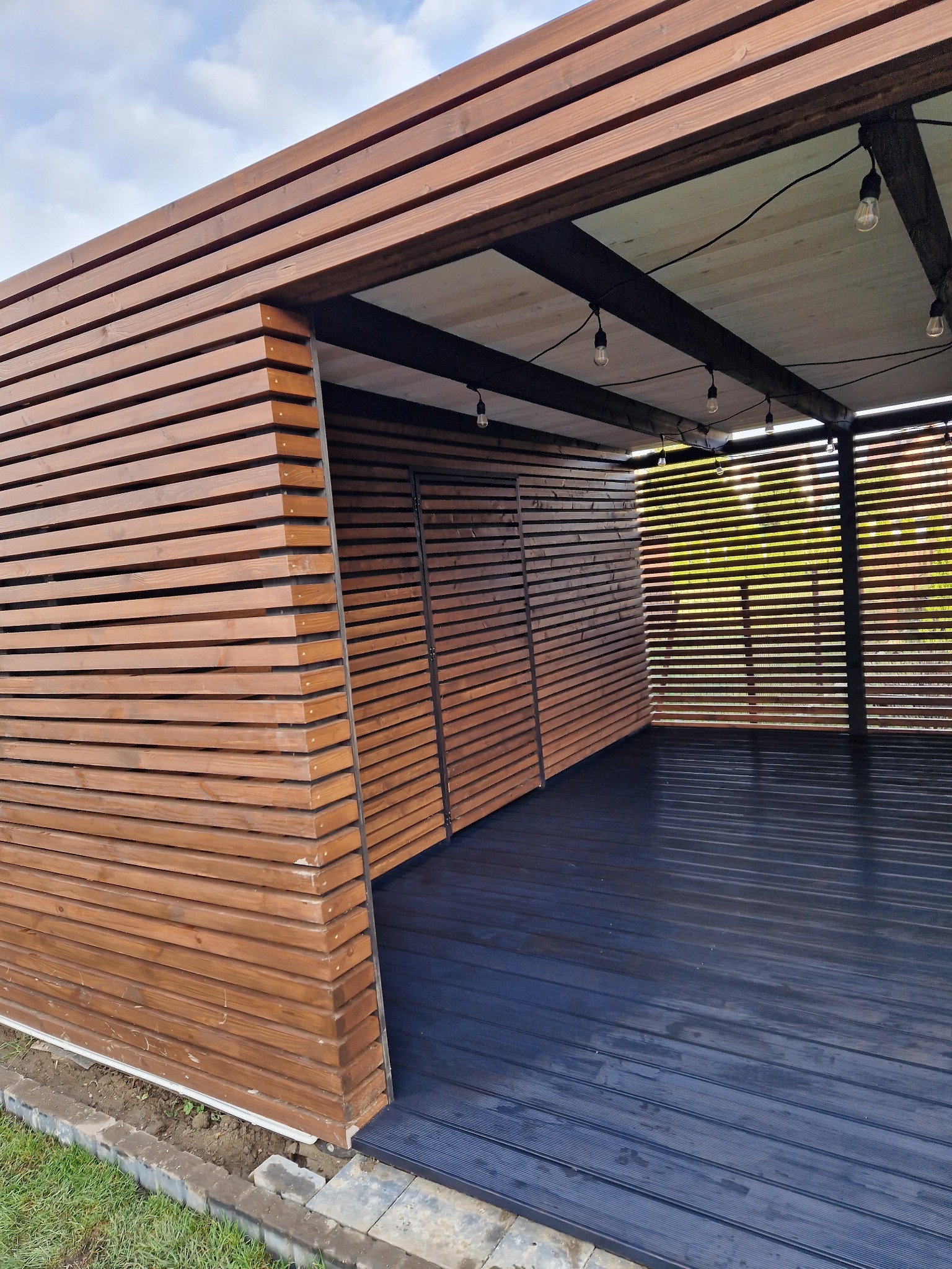 Nowoczesna zabudowa balkonu z drewnianych lameli w odcieniach brązu, z czarną podłogą i dekoracyjnym oświetleniem w postaci wiszących żarówek.