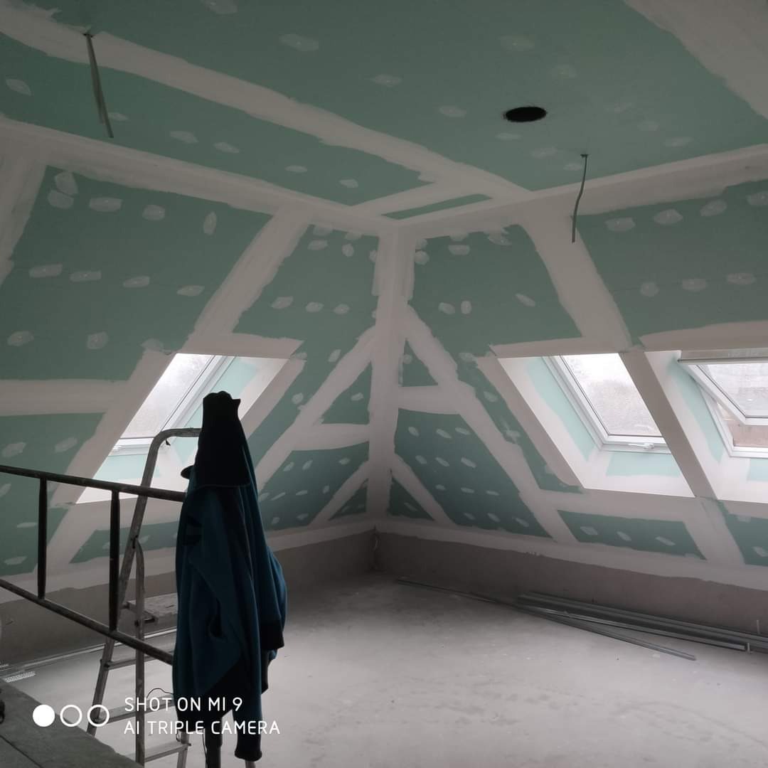 Poddasze w trakcie prac wykończeniowych: płyty karton-gips na skosach i suficie, z widocznymi łączeniami zaszpachlowanymi na biało, dwa okna dachowe, drabina z powieszonym ubraniem w tle.