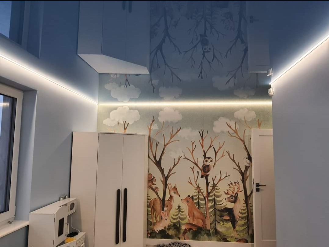 Pokój dziecięcy z błyszczącym, niebieskim sufitem podwieszanym z motywem drzew i zwierząt, podświetlony listwami LED, ściany z tapetą w leśny wzór, białe meble.