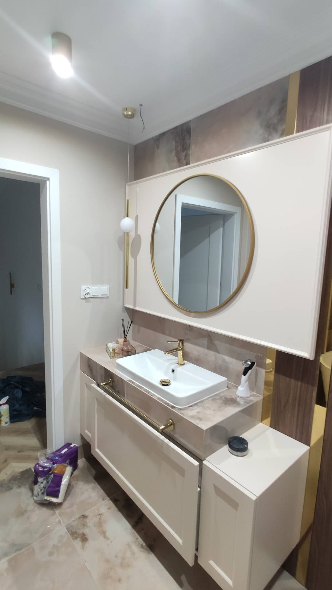 Nowoczesna łazienka z beżową szafką pod umywalką z marmurowym blatem, okrągłym lustrem w złotej ramie i złotą armaturą. Widoczne są również dekoracyjne elementy oświetleniowe oraz częściowo otwarte...