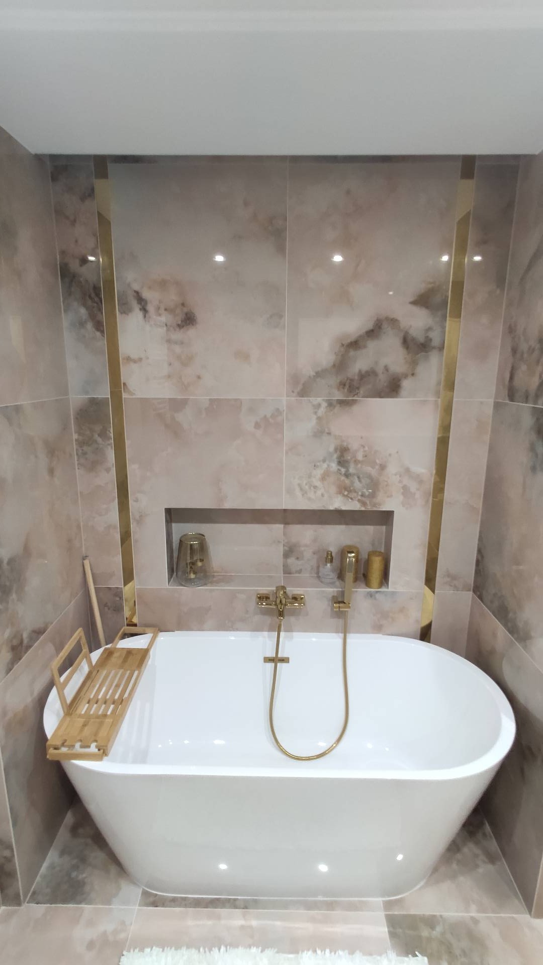 Nowoczesna łazienka z owalną białą wanną, złotą armaturą i marmurkowymi płytkami z akcentami w kolorze złota, półka w ścianie z dekoracjami, drewniana półka na wannę.