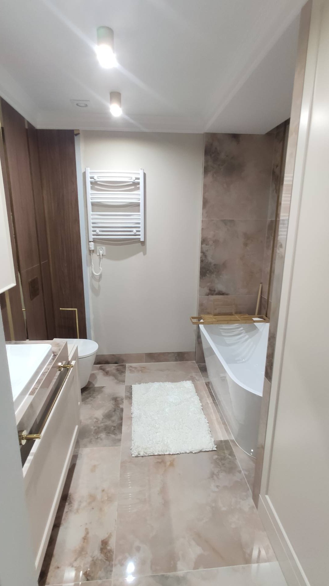 Nowoczesna łazienka z wanną, grzejnikiem drabinkowym i marmurową podłogą, oświetlona punktowo, z widocznymi detalami w kolorze złotym.