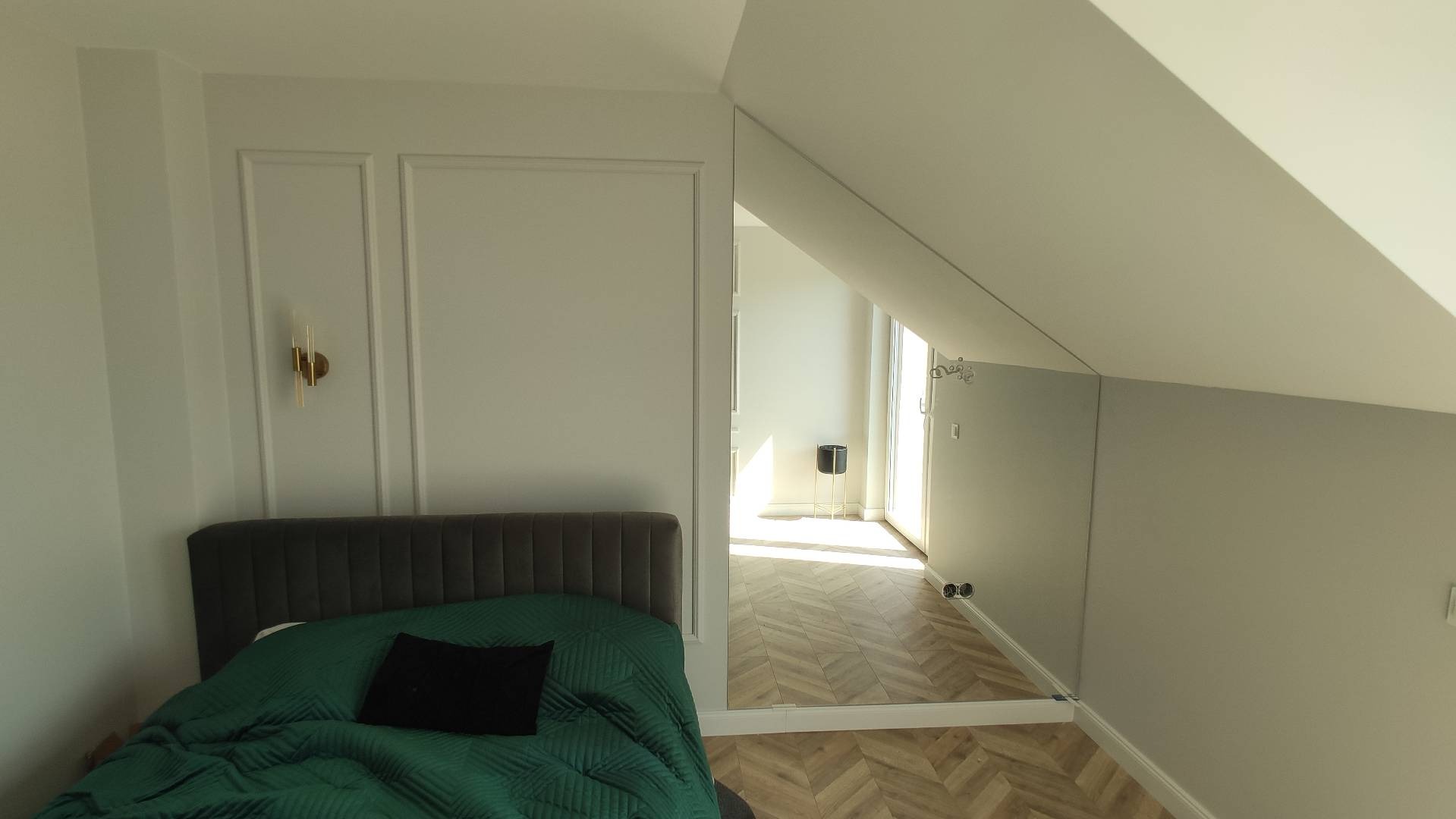 Pokój na poddaszu z łóżkiem z zieloną narzutą i czarną poduszką, widok na korytarz z parkietem ułożonym w jodełkę, ściany w kolorze złamanej bieli z dekoracyjnymi listwami i złotym kinkietem.