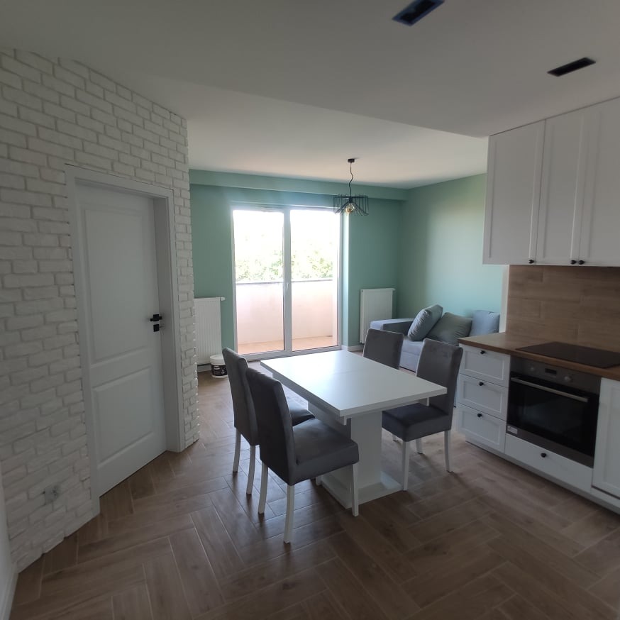 Jasne wnętrze mieszkania po remoncie: biała cegła na ścianie, podłoga ułożona w jodełkę, biały stół z szarymi krzesłami, aneks kuchenny z białymi szafkami i drewnianym blatem, widok na balkon...