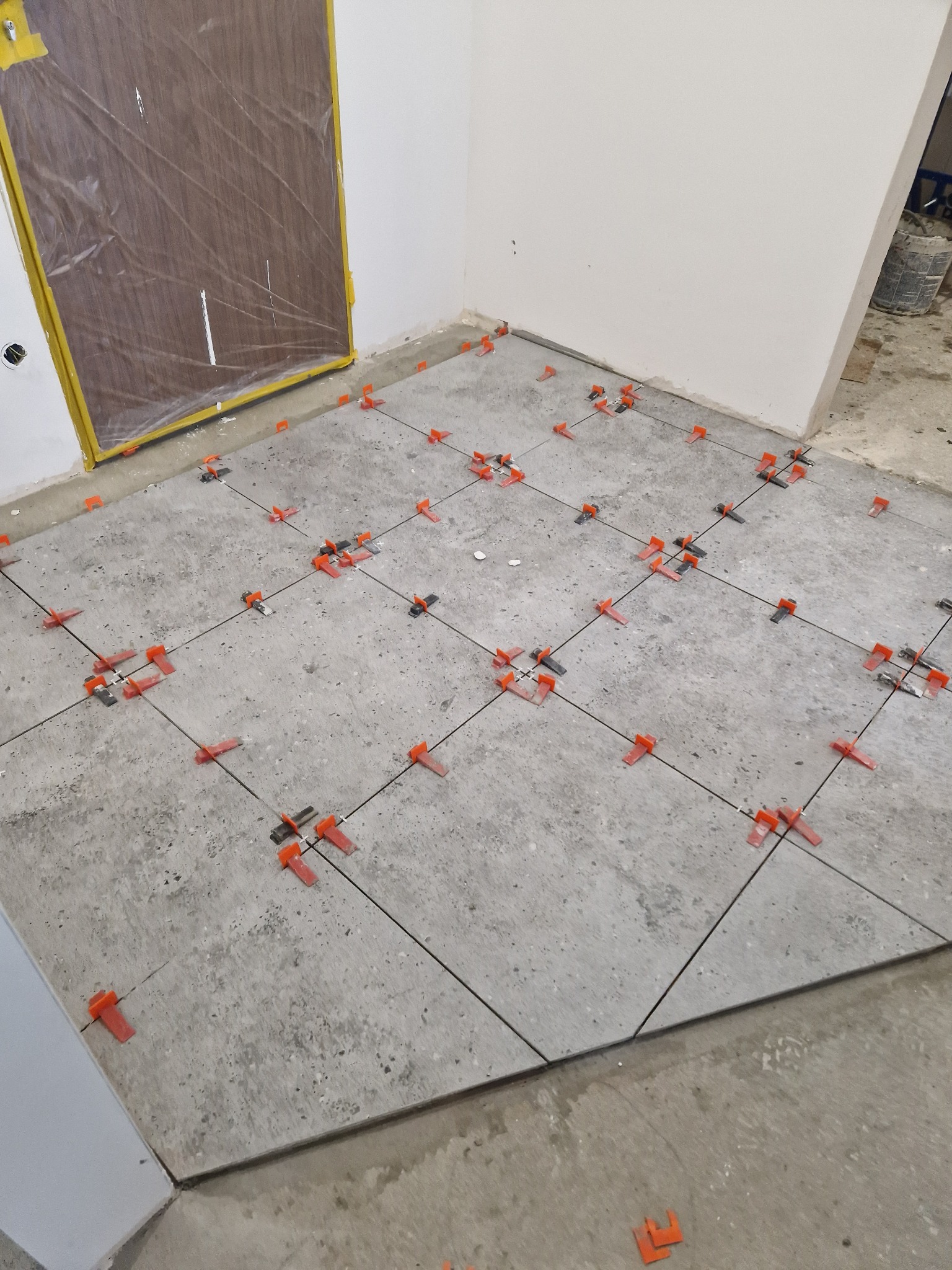 Układanie szarych płytek podłogowych z systemem poziomowania, widoczne krzyżyki dystansowe i sznurek wyznaczający linie, zabezpieczone drzwi folią ochronną.
