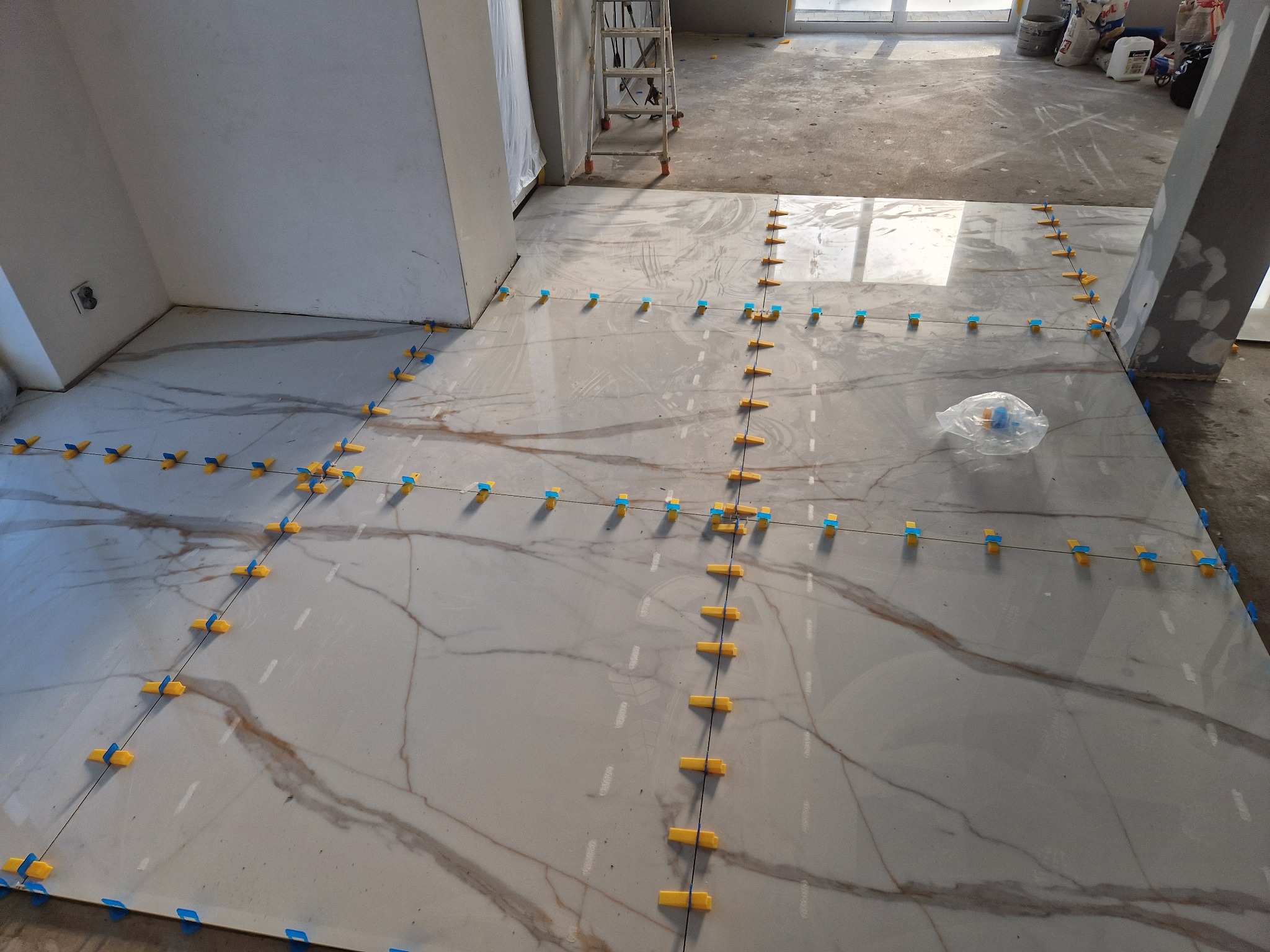 Układanie płytek podłogowych z wzorem marmuru, z użyciem żółto-niebieskich klipsów poziomujących i czarnej żyłki, na tle surowego stanu wykończenia wnętrza.
