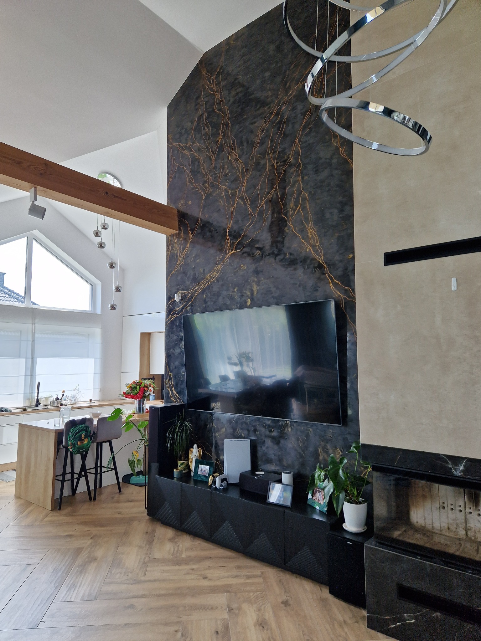 Nowoczesny salon z otwartą kuchnią, ściana akcentująca z efektem marmuru i złotymi żyłkami, telewizor, kominek z czarnego marmuru, parkiet w jodełkę, designerska lampa wisząca z metalowych okręgów.