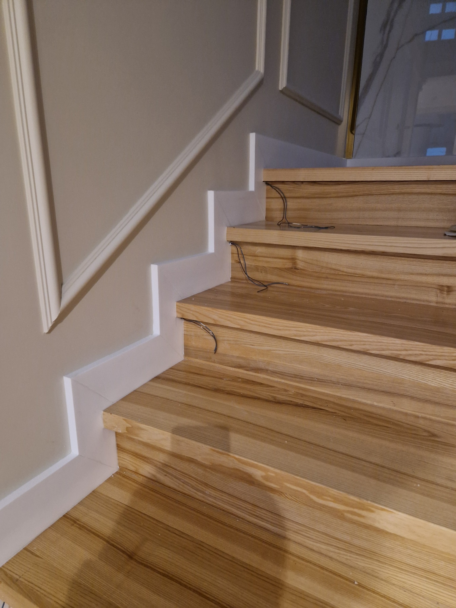 Wykończone drewniane schody z białymi listwami przypodłogowymi i dekoracyjną sztukaterią na ścianie, widoczne wystające kable instalacyjne na stopniach.