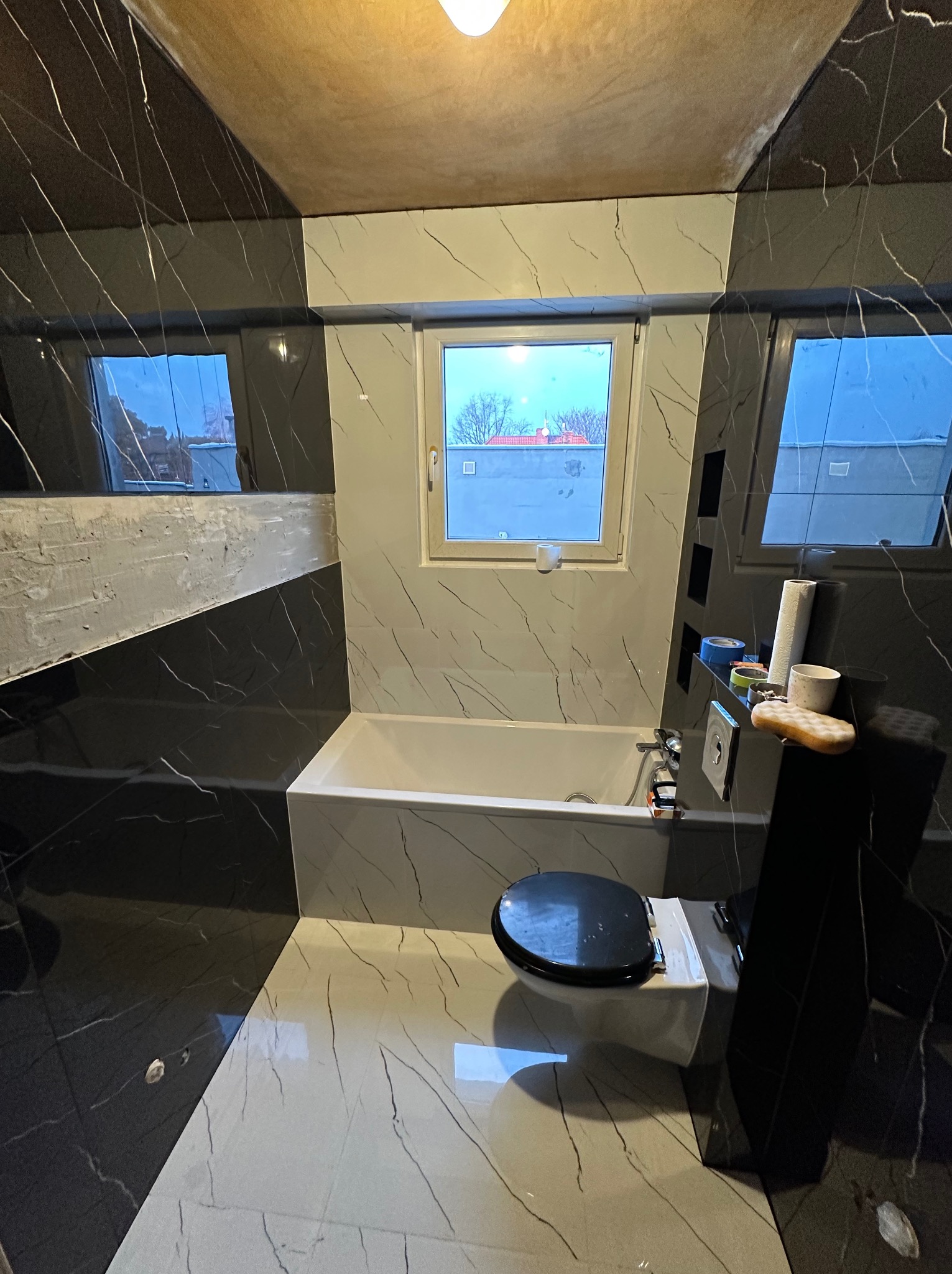 Łazienka w trakcie wykańczania, z czarno-białą marmurkową glazurą na ścianach i podłodze, białą wanną i wiszącą toaletą z czarną deską. Na półce przy wannie widoczne akcesoria łazienkowe i narzędzia.