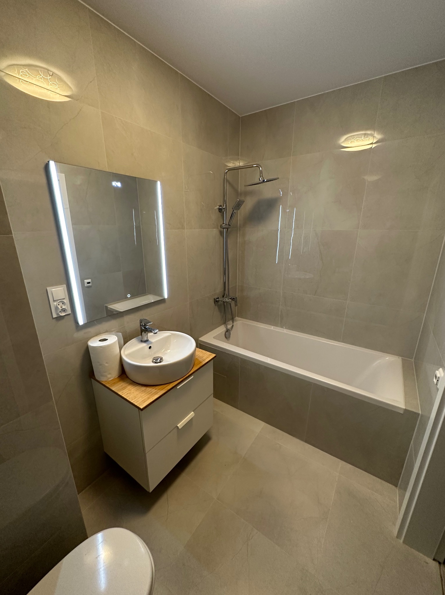 Nowoczesna łazienka w odcieniach szarości z podświetlanym lustrem, okrągłą umywalką na szafce z blatem imitującym drewno i białą wanną z zamontowaną baterią prysznicową.