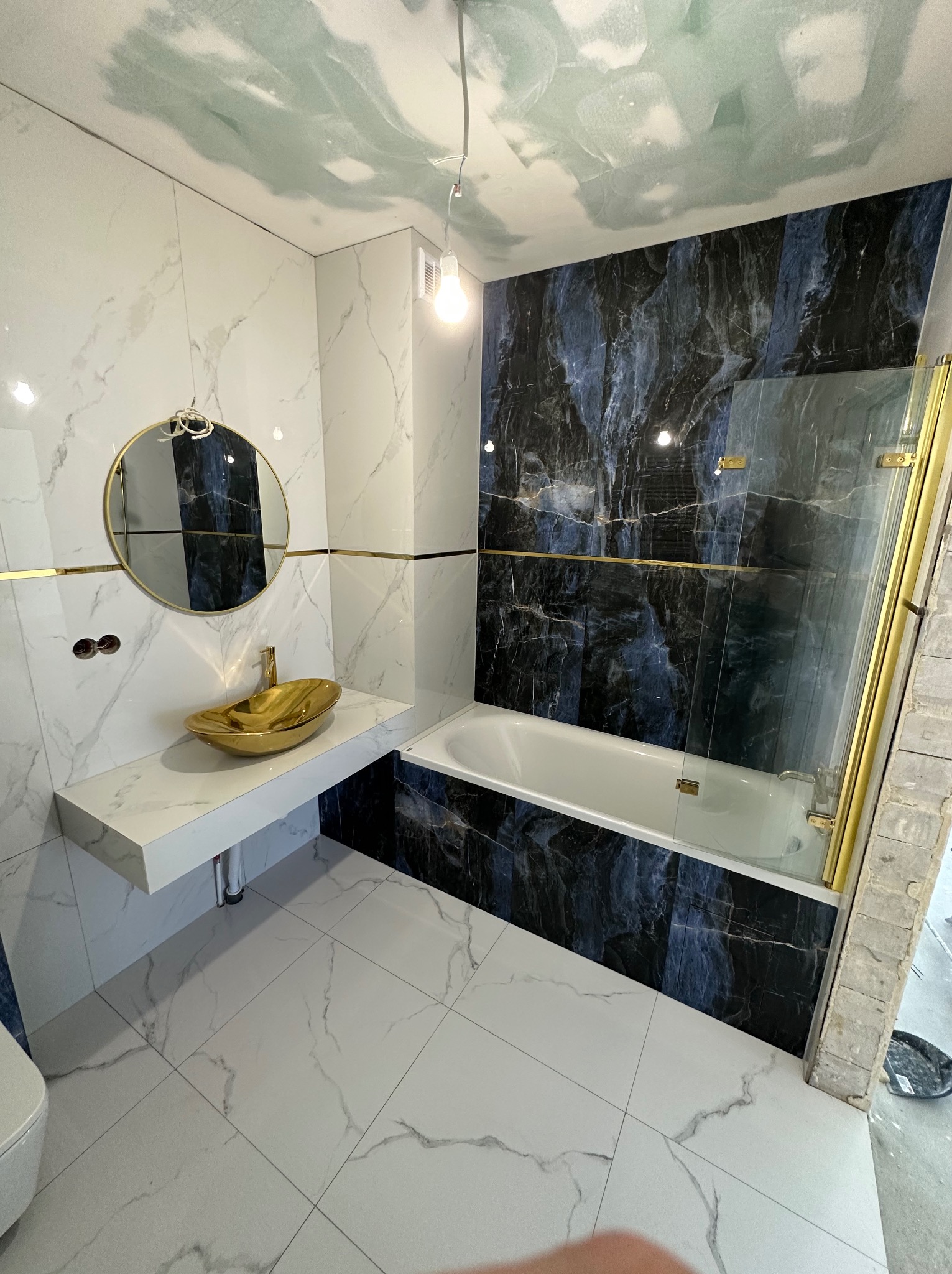 Nowoczesna łazienka w trakcie wykańczania: białe płytki imitujące marmur na podłodze i ścianach, złota umywalka na białym blacie, czarne płytki imitujące marmur przy wannie, szklany parawan...
