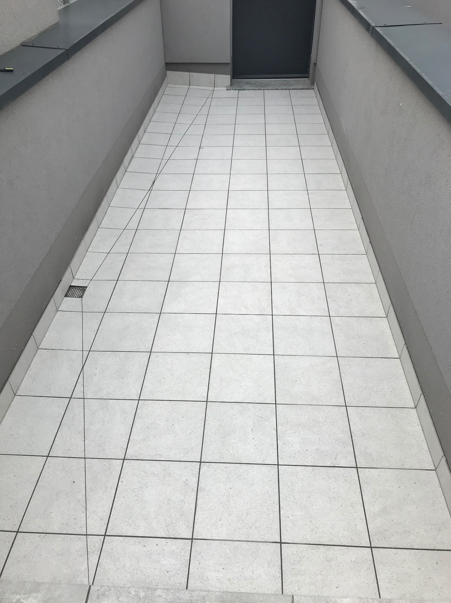 Wyłożony jasnymi płytkami taras z odpływem liniowym przy ścianie, widok z góry, szare ściany i metalowe obramowanie.