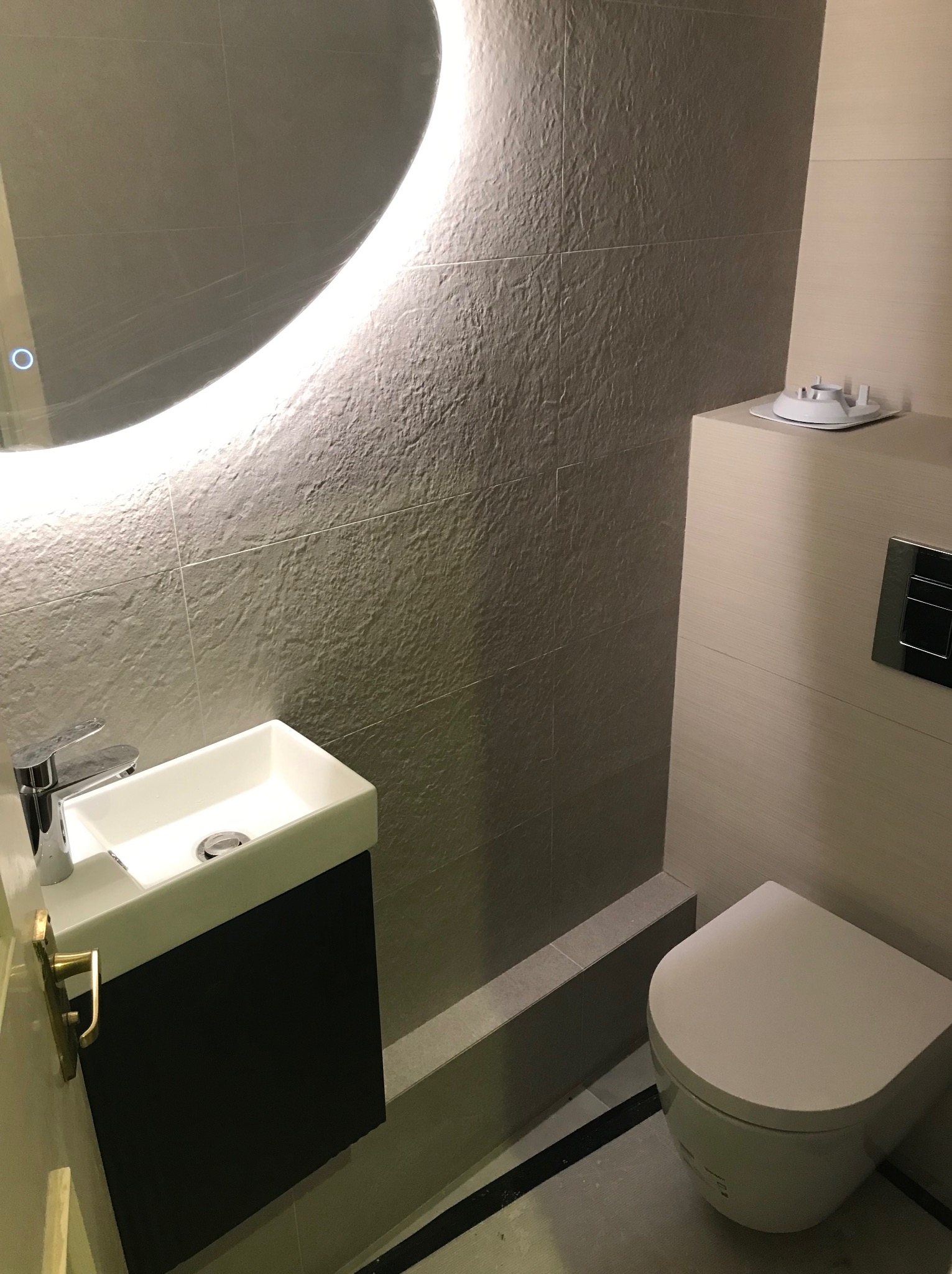Nowoczesna łazienka z umywalką nablatową na czarnej szafce, podświetlanym lustrem i podwieszaną toaletą z szarą, kamienną okładziną ścienną.