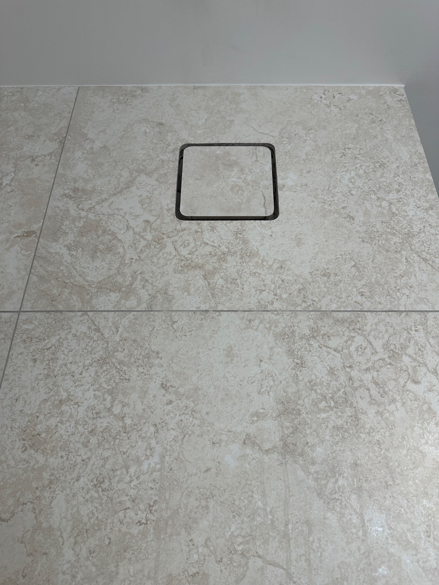Jasne, marmurkowe płytki w łazience z kwadratowym odpływem liniowym w podłodze, wykończonym czarną ramką.