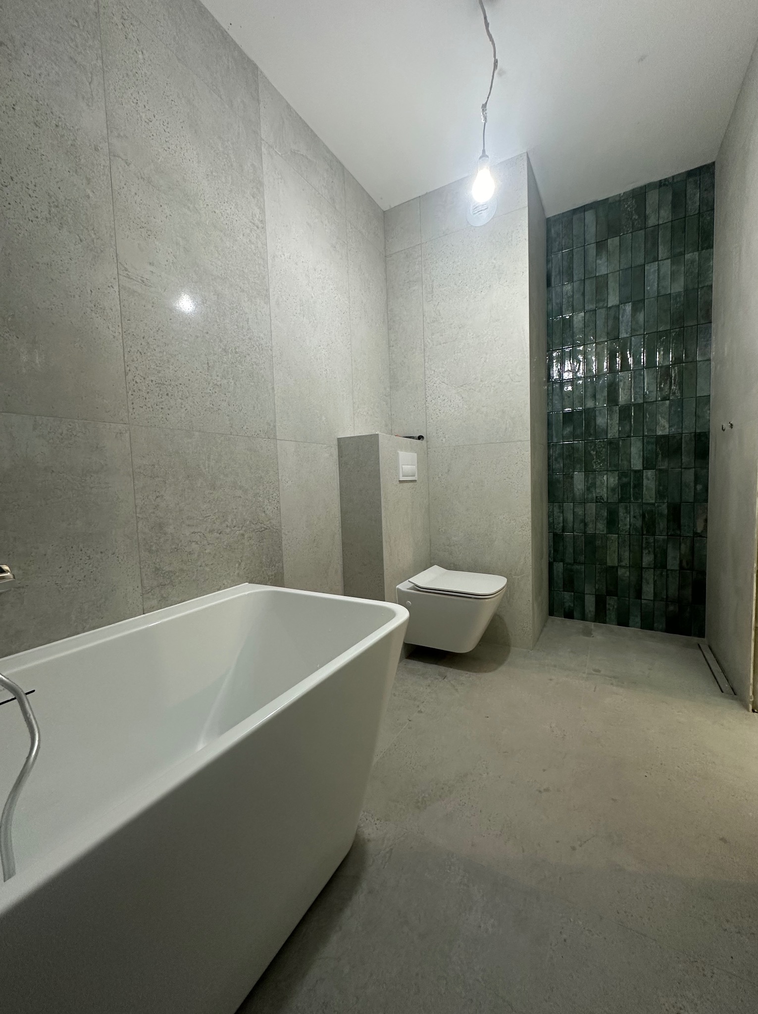 Nowoczesna łazienka w trakcie wykańczania: biała wanna prostokątna, podwieszana toaleta i ściana z ciemnozielonych, pionowo ułożonych płytek, szare płytki na ścianach i podłodze, surowy stan podłoża.