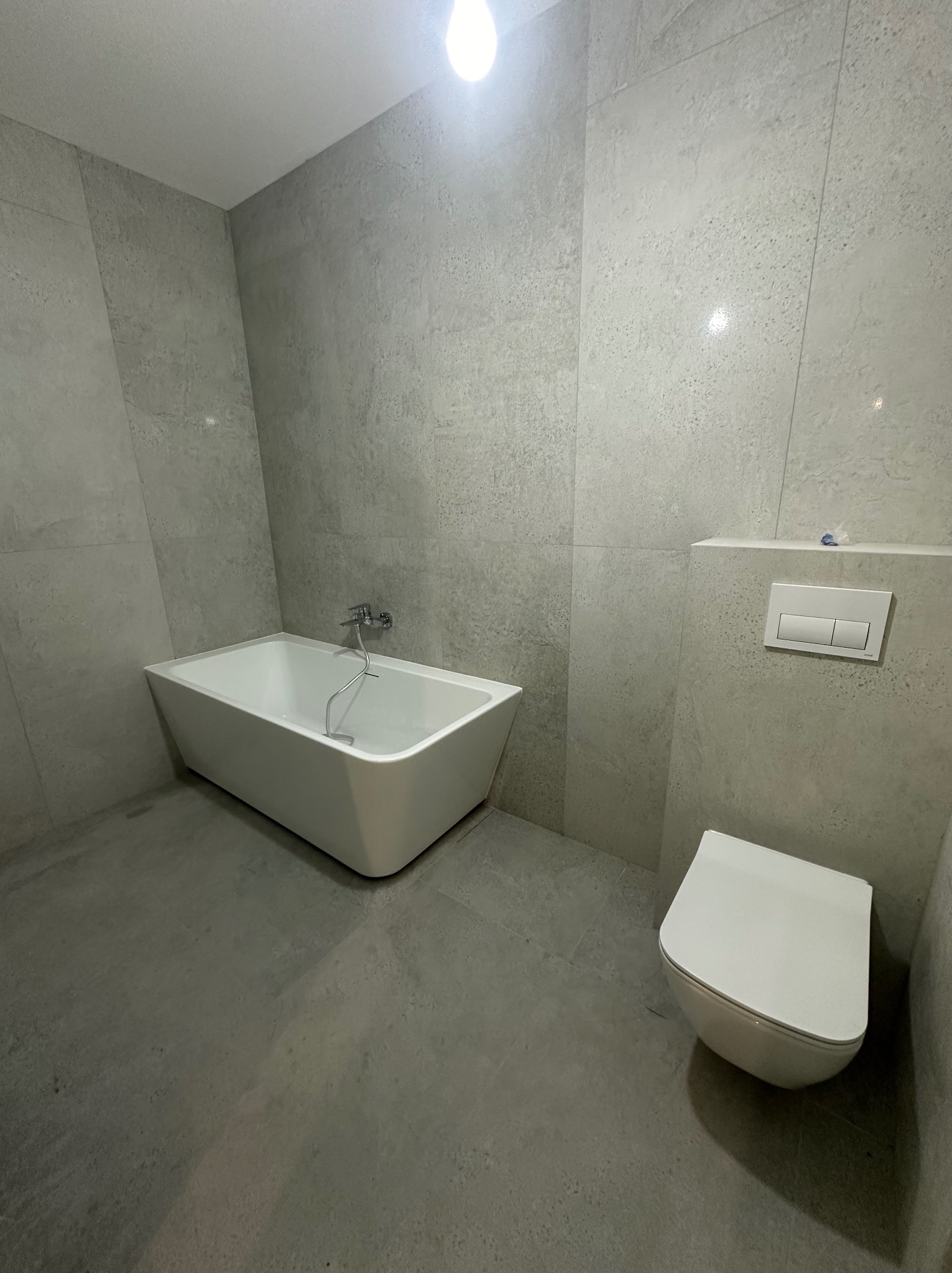 Nowoczesna łazienka z wanną wolnostojącą i podwieszaną toaletą, wykończona szarymi płytkami imitującymi kamień, oświetlona górnym światłem.