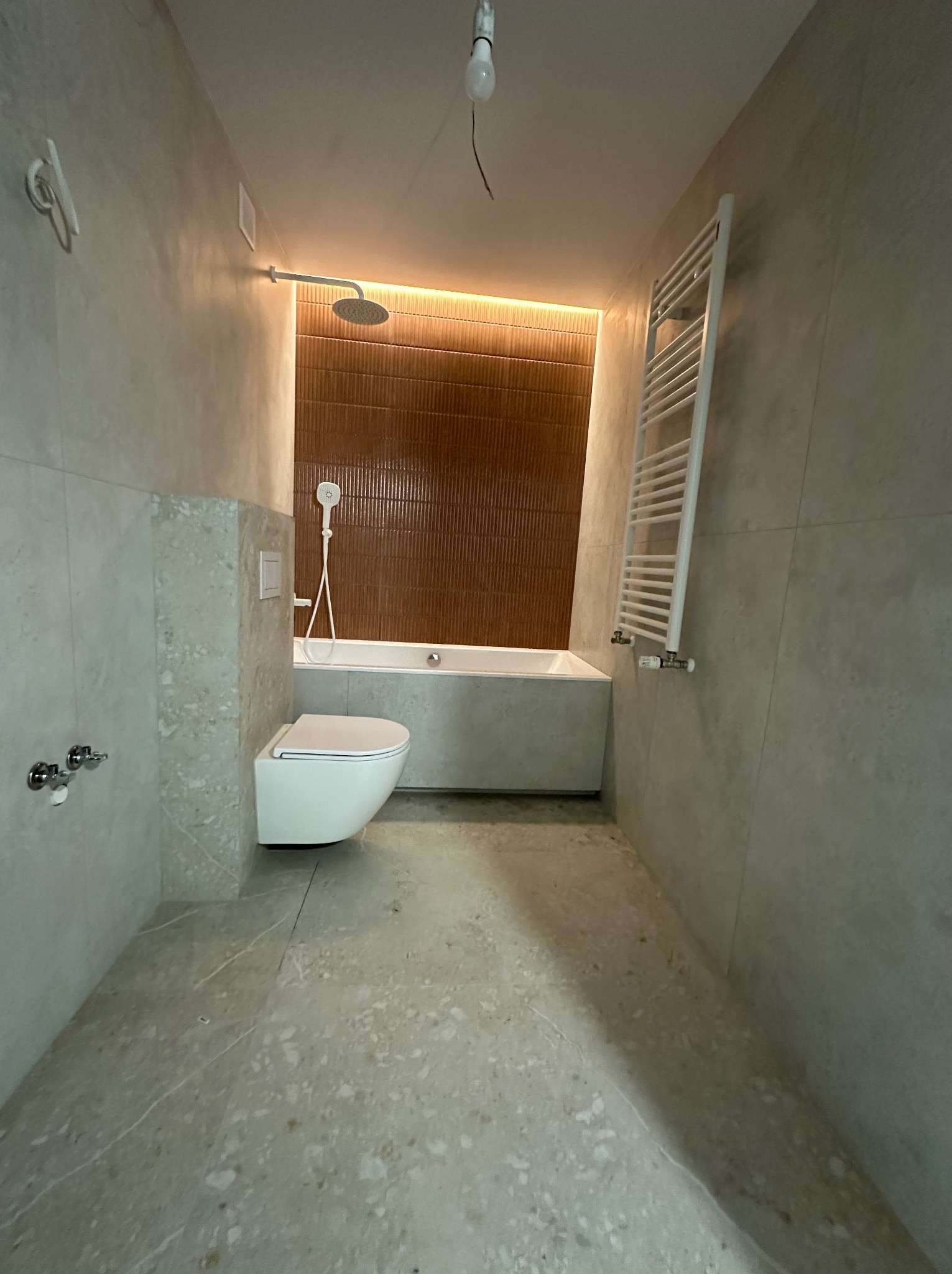 Nowoczesna łazienka z beżowymi płytkami, podświetlaną ścianą z brązową teksturą, białą wanną, toaletą i grzejnikiem drabinkowym. Widoczna surowa żarówka na suficie.