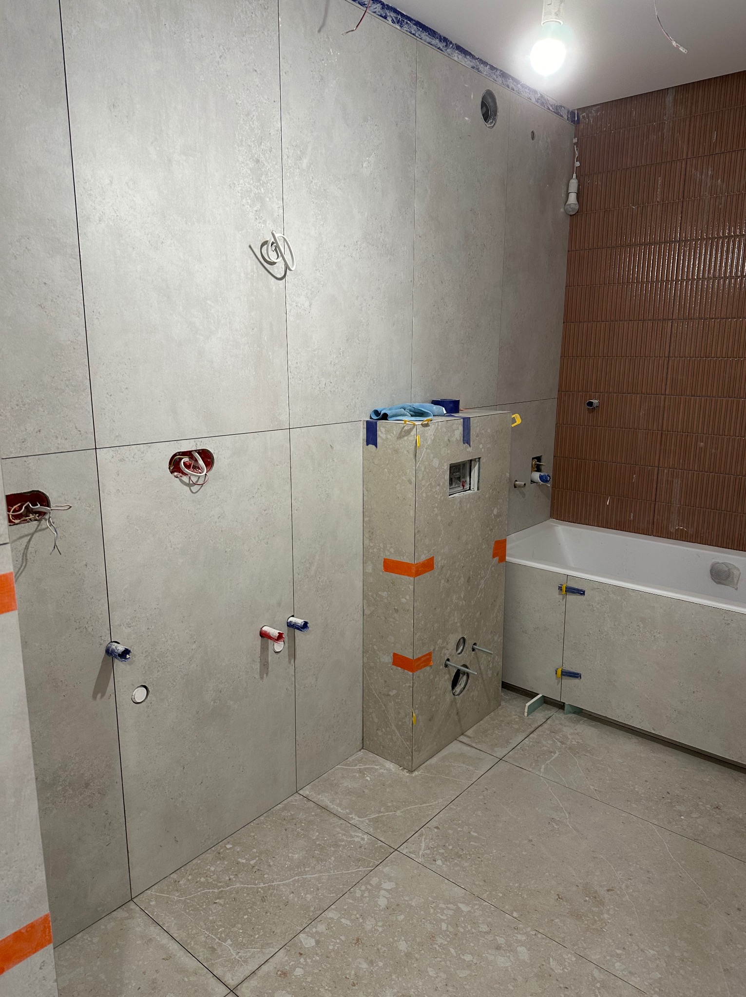 Łazienka w trakcie prac wykończeniowych: ułożone płytki na ścianach i podłodze w odcieniach szarości, widoczne instalacje hydrauliczne i elektryczne, zabudowa WC z pomarańczową taśmą, fragment...