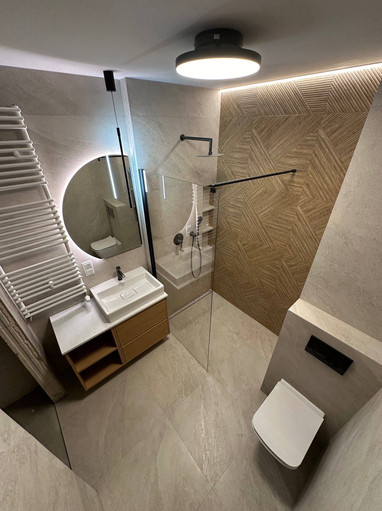 Nowoczesna łazienka z kabiną prysznicową typu walk-in, umywalką nablatową na drewnianej szafce, podświetlanym lustrem i grzejnikiem drabinkowym, wykończona płytkami w odcieniach beżu i brązu...