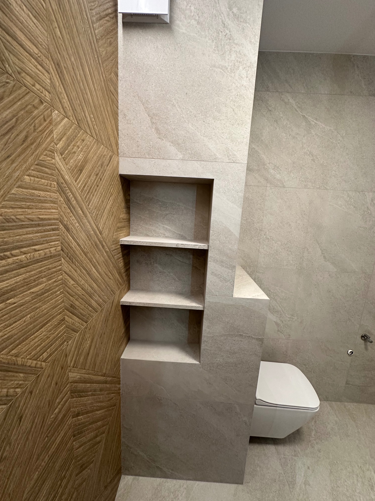 Nowoczesna łazienka z drewnopodobną ścianą w geometryczne wzory, wbudowane półki z szarej płytki oraz podwieszana toaleta.