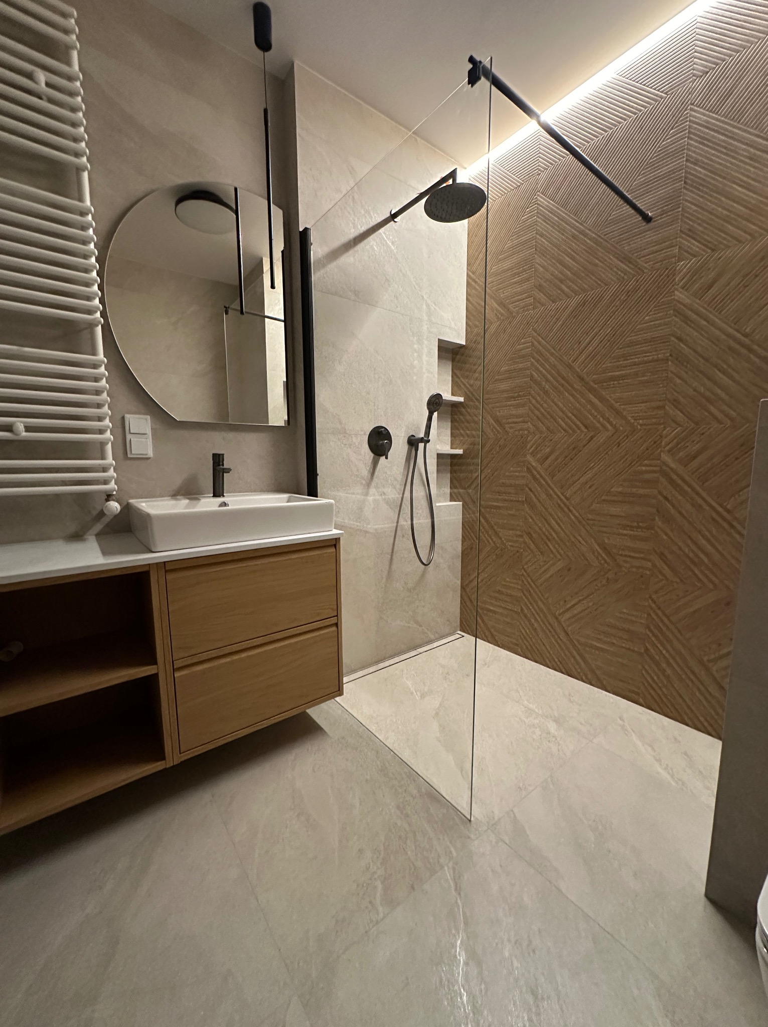 Nowoczesna łazienka z kabiną prysznicową typu walk-in, umywalką nablatową na drewnianej szafce i dekoracyjną ścianą z płytek o geometrycznym wzorze w odcieniach brązu.