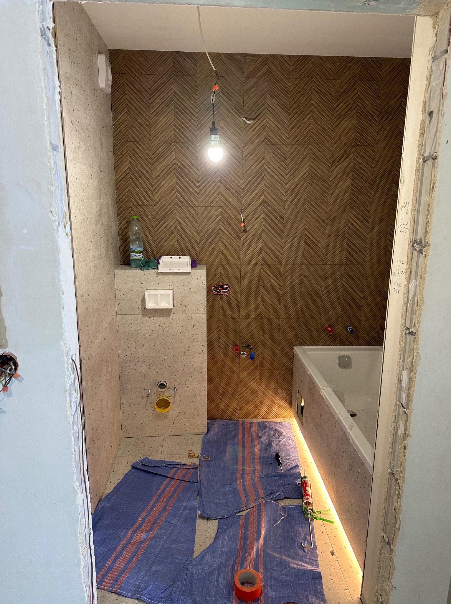 Wnętrze łazienki w trakcie prac wykończeniowych, widoczne płytki imitujące drewno ułożone w jodełkę na ścianie, częściowo obudowana wanna z podświetleniem LED, zabezpieczona podłoga płachtą...