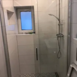 Nowoczesna kabina prysznicowa z deszczownicą i oknem, wykończona białymi płytkami i wzorzystą podłogą, widok na częściowo widoczną toaletę.