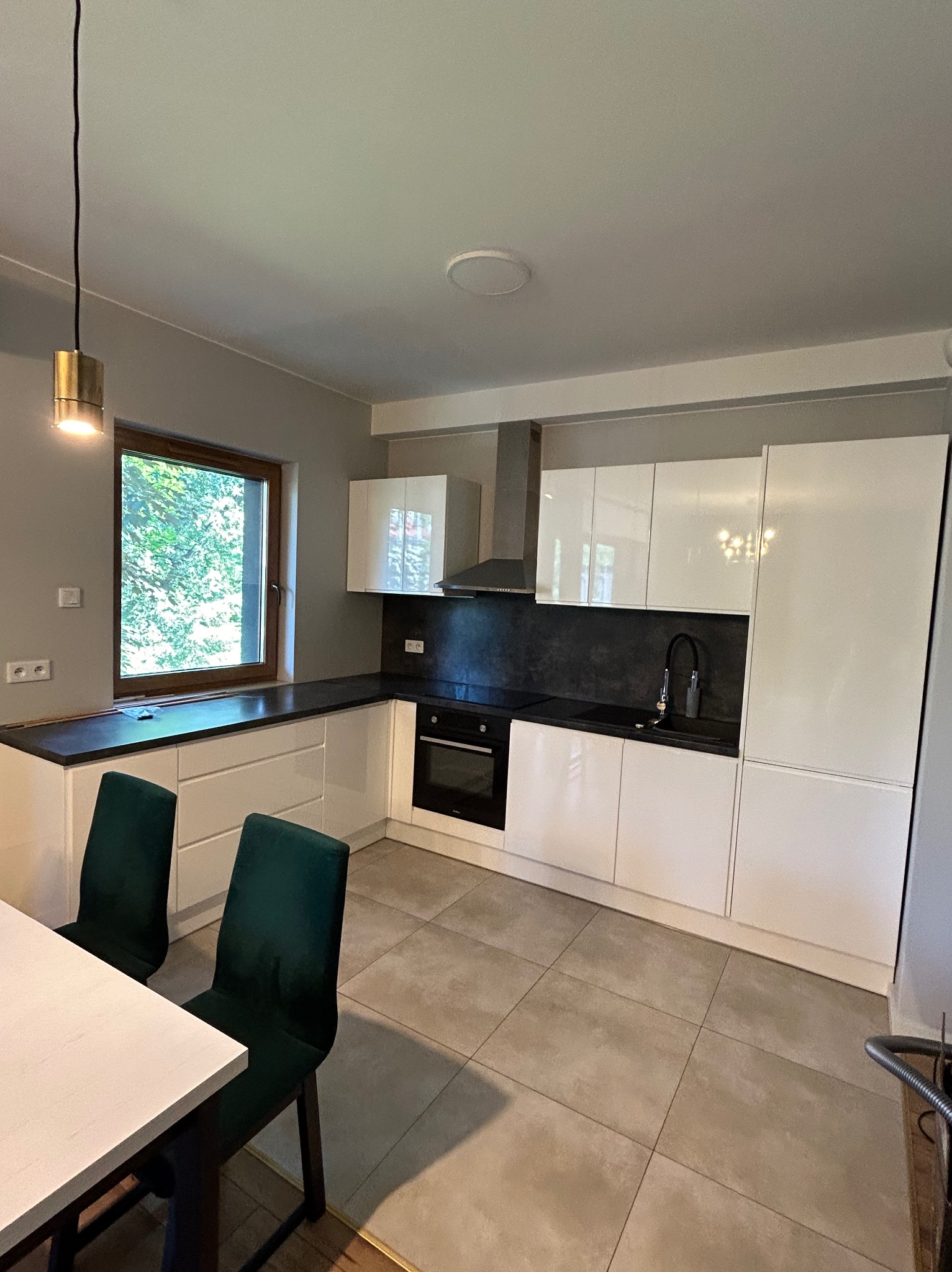 Nowoczesna kuchnia z białymi szafkami, czarnym blatem i płytkami podłogowymi w odcieniach szarości, oświetlona złotą lampą wiszącą nad stołem z zielonymi krzesłami.