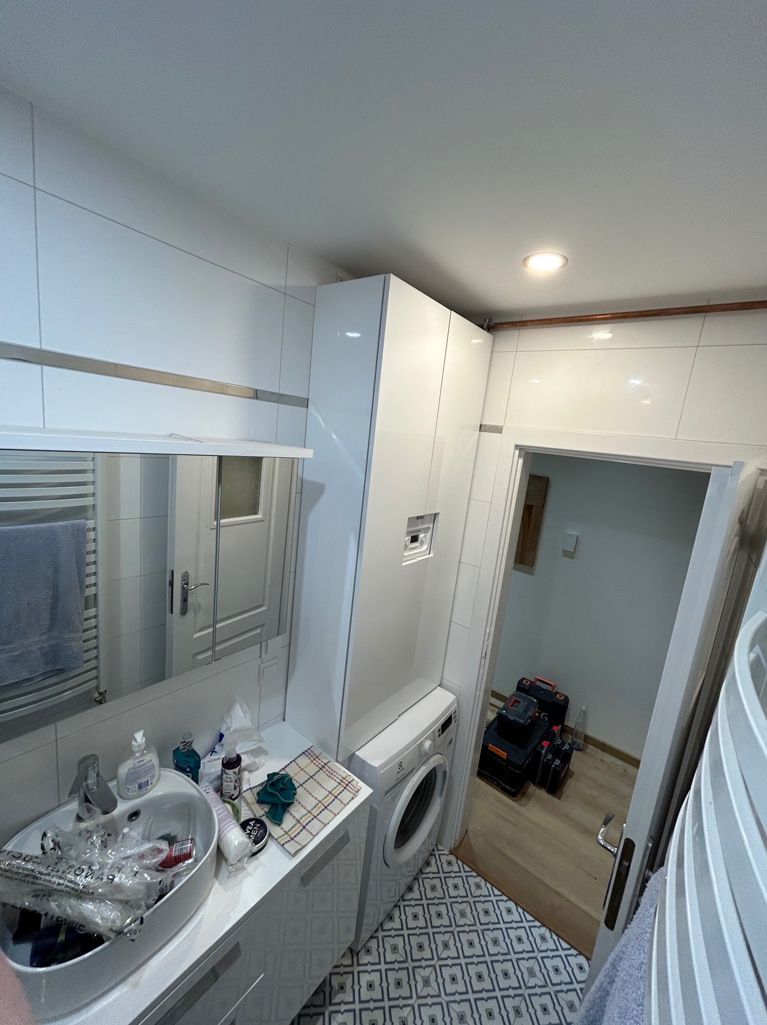 Wnętrze łazienki w trakcie prac, widoczna umywalka z akcesoriami, pralka, zabudowa meblowa, narzędzia w skrzynkach stojące w przejściu do innego pomieszczenia.