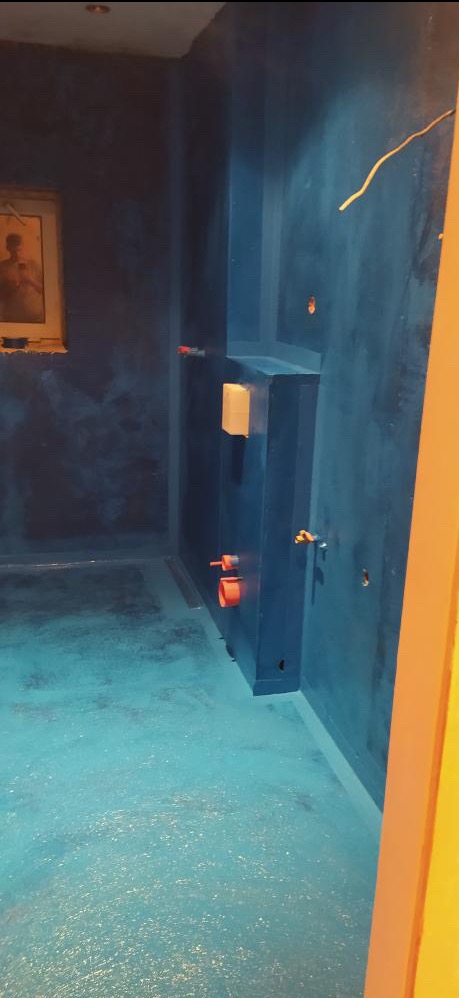 Niebieska, błyszcząca łazienka w trakcie prac wykończeniowych, widoczne wystające rury i przewody elektryczne, obraz w tle, oświetlenie sufitowe.