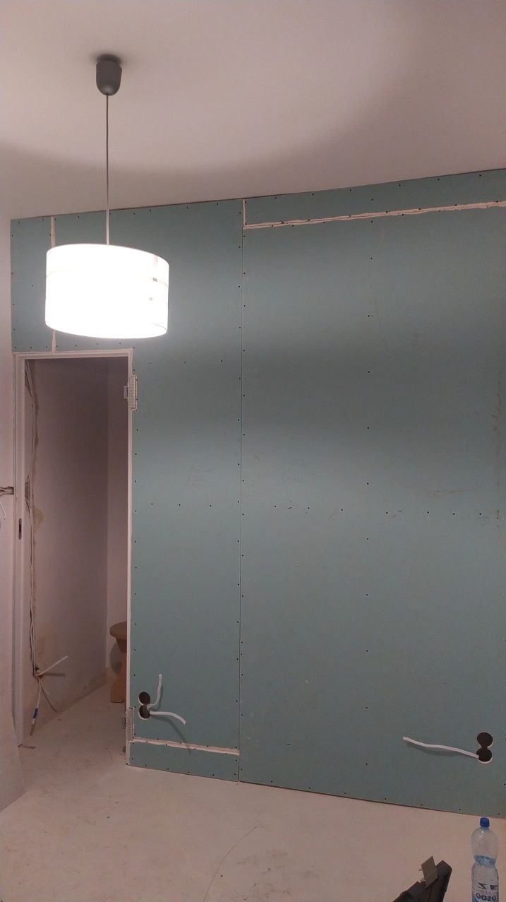 Pokój w trakcie remontu: ściany pokryte płytami gipsowo-kartonowymi, widoczne kable elektryczne wystające ze ściany, surowa podłoga, wisząca lampa z abażurem, butelka wody na podłodze.