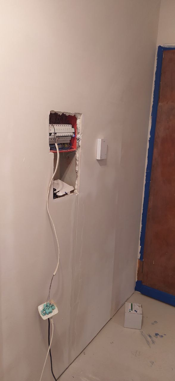 Ściana w trakcie przygotowania do malowania z widoczną otwartą skrzynką elektryczną i wystającymi przewodami, zabezpieczone drzwi taśmą malarską.
