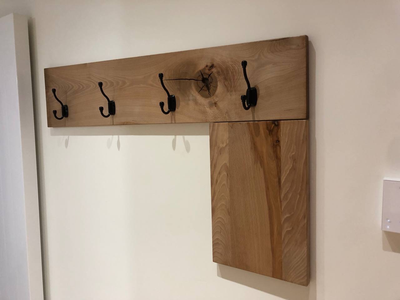 Drewniany wieszak z pięcioma czarnymi haczykami, zamontowany na ścianie, z widocznym sękiem w drewnie i dodatkową półką z boku.