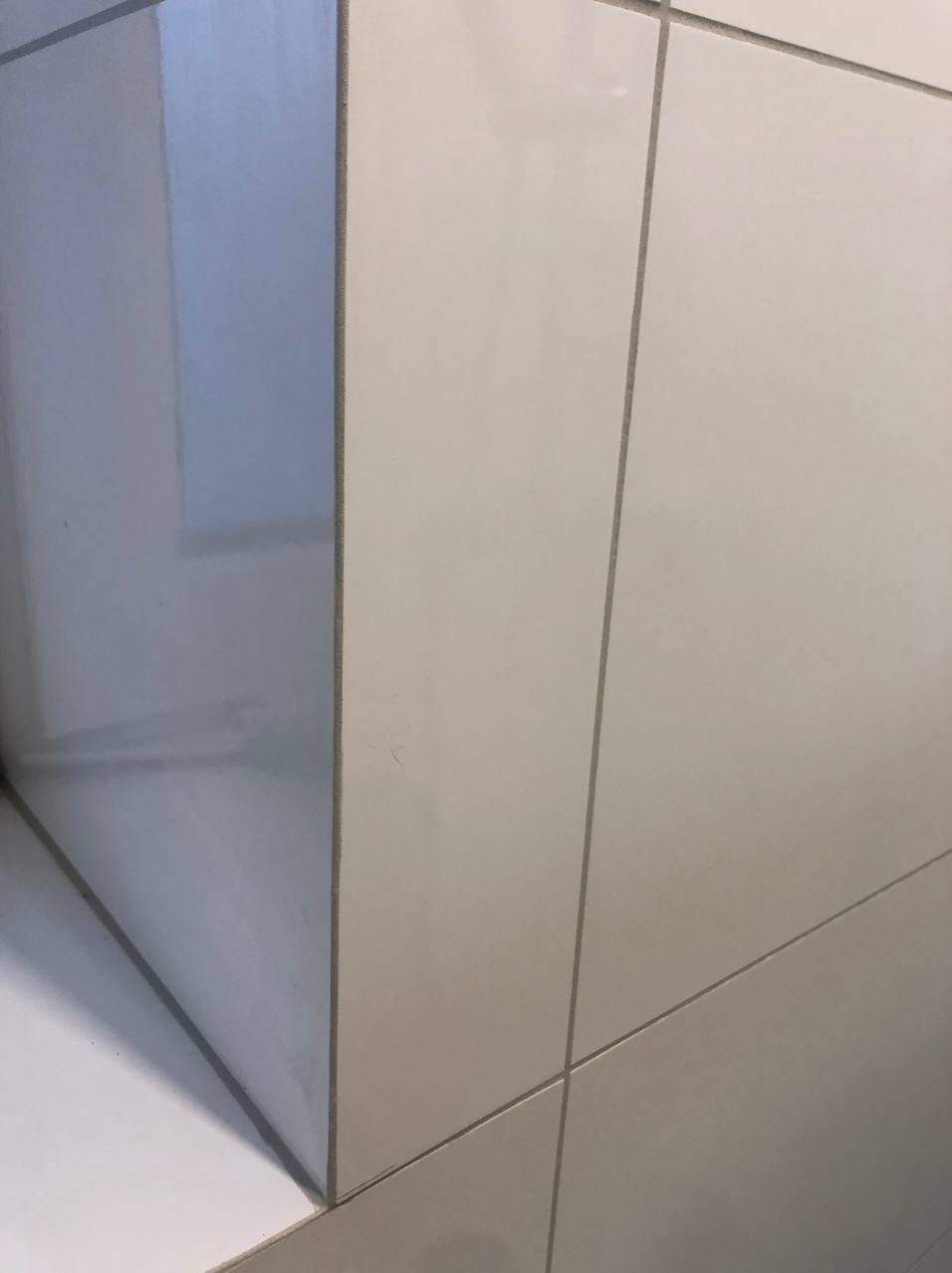 Fragment ściany wyłożonej jasnymi, błyszczącymi płytkami ceramicznymi z szarą fugą, widoczny narożnik ściany z odbiciem światła.