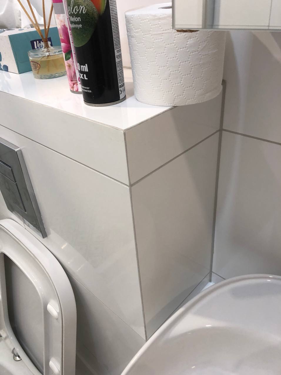 Biała, minimalistyczna łazienka z toaletą, umywalką i szafką z akcesoriami, w tym odświeżacz powietrza i papier toaletowy.