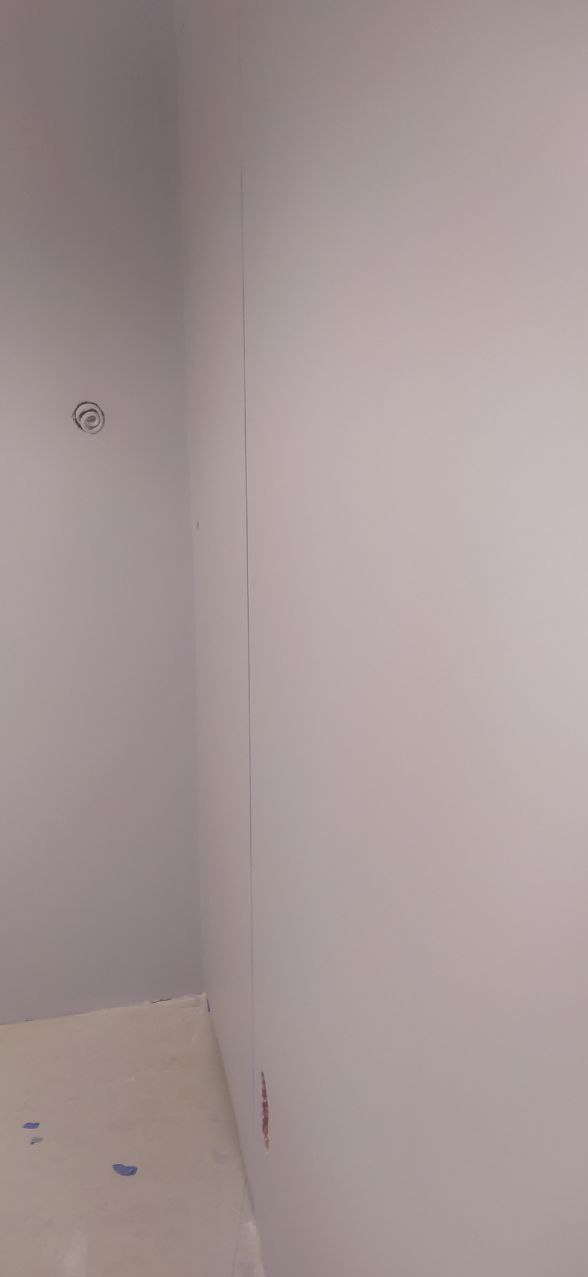 Świeżo pomalowana ściana w odcieniach szarości, widoczna puszka elektryczna bez osprzętu oraz ślady po usuniętej taśmie malarskiej na podłodze i ścianie.