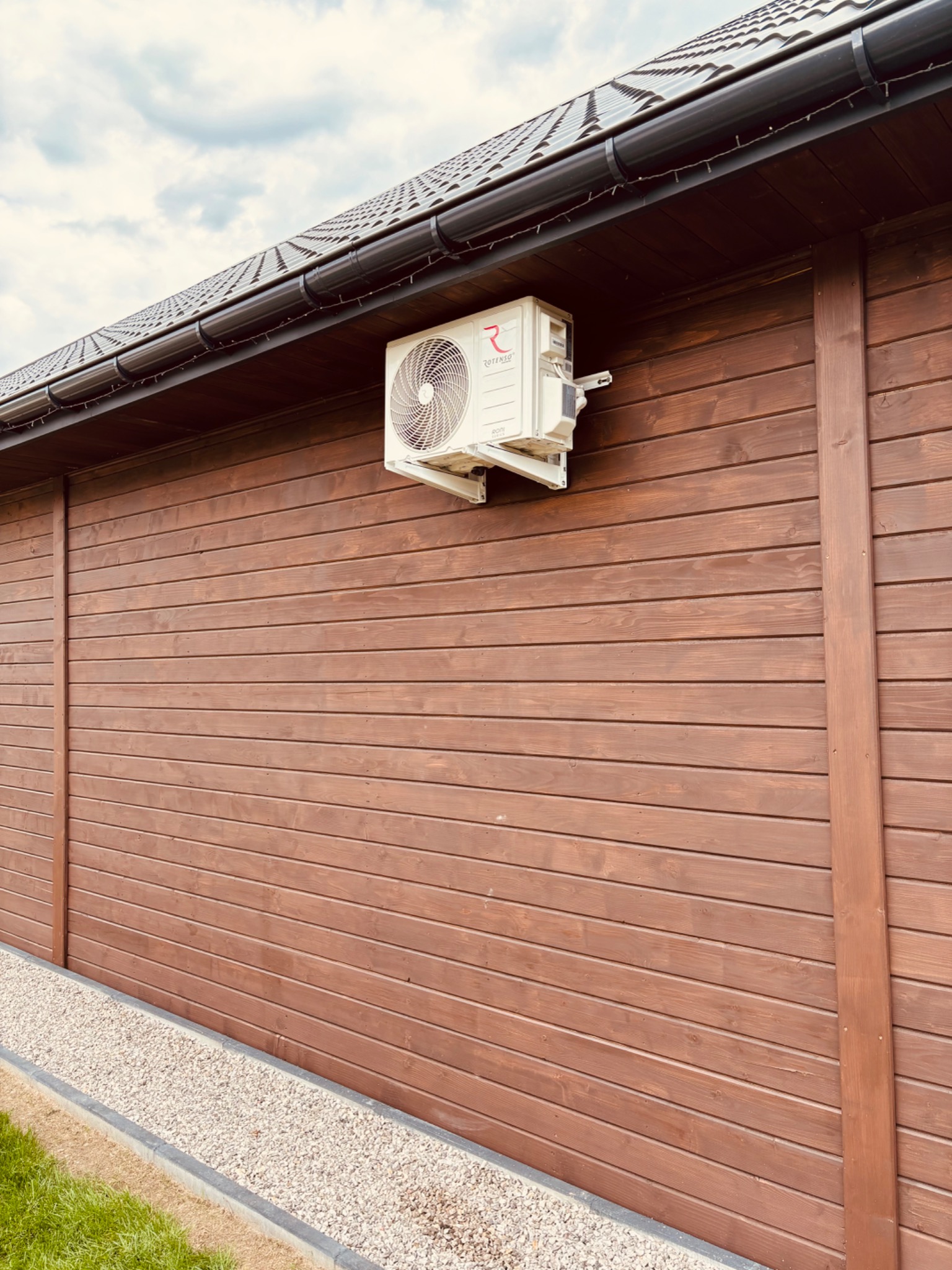 Klimatyzator marki Rotenso zamontowany na brązowej, drewnianej ścianie budynku z widocznym dachem i rynną. Obok ściany znajduje się kamienny chodnik.